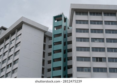White buildings flank a turquoise elevator shaft. Overcast sky looms above the structures. Windows align in repeating geometric patterns. Construction or renovation appears incomplete
