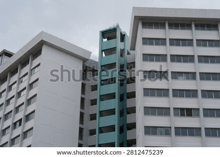 White buildings flank a turquoise elevator shaft. Overcast sky looms above the structures. Windows align in repeating geometric patterns. Construction or renovation appears incomplete