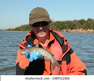 A white bullhead catfish being held by a smiling woman in a baseball cap and black and red coat on a river bay on a bright sunny day