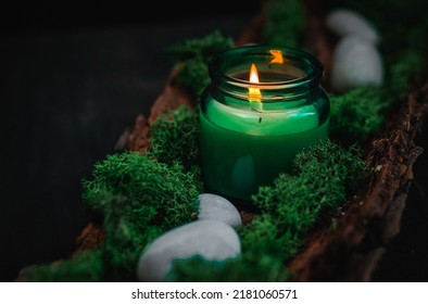 A white burning candle in a glass jar with green moss and gray stones in the bark of a tree lie on a black background, close-up side view. Esoteric concept, dark style. - Powered by Shutterstock