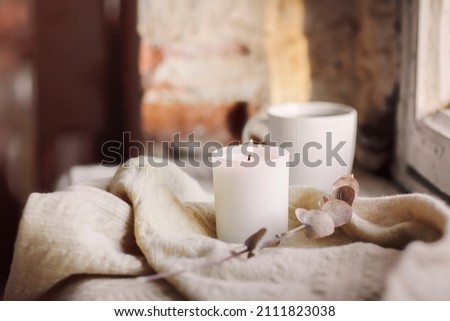 White burning candle, woolen scarf or plaid and cup of hot tea on windowsill inside old house, selective focus. Creating cozy and inviting atmosphere by adding candles to home decor
