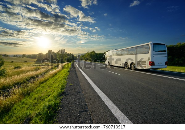 White bus traveling on the asphalt road in a rural\
landscape at sunset