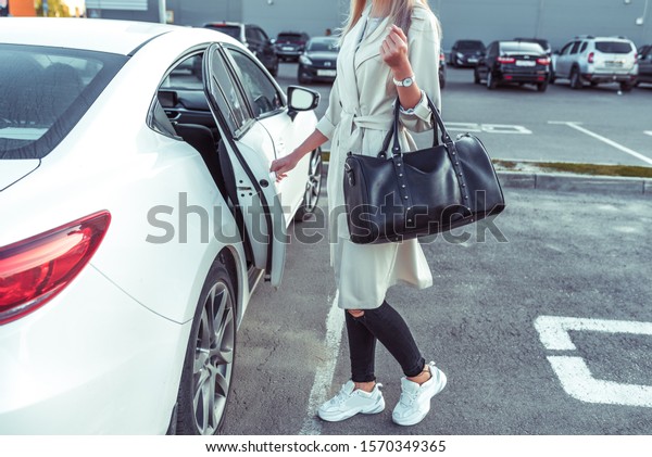 White business class sedan, woman opens door of\
taxi, car on call, car sharing. In summer in city. White cloak and\
leather bag. Parking at\
mall