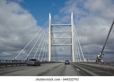 White cable-stayed bridge, view from the road. Asphalt motorway with cars against a blue sky with clouds.