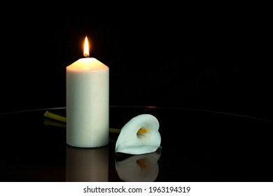 WHITE CALLA FLOWER NEXT TO A LIGHTED CANDLE ON DARK BACKGROUND. ALL SOULS DAY, DEATH, DECEASE, PRAYER, MEMORIAL DAY, MOURNING, GRAVE, CEMETERY, BURIAL, CONDOLENCE AND FUNERAL CONCEPT. COPY SPACE.