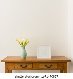 White Calla Lilies In Teal Vase With Blank Square Picture Frame On Oak Side Table Against White Wall