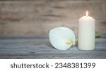 WHITE CALLA LILY FLOWER NEXT TO A LIGHTED CANDLE ON WOODEN BACKGROUND. CREMATION, BURIAL, FUNERAL, CONDOLENCE CARD. COPY SPACE.