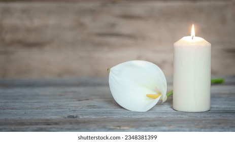 WHITE CALLA LILY FLOWER NEXT TO A LIGHTED CANDLE ON WOODEN BACKGROUND. CREMATION, BURIAL, FUNERAL, CONDOLENCE CARD. COPY SPACE.