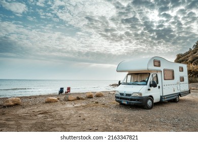 White camper parked in front of seaside, two chairs on pebbles beach, one man sitting and relaxing on chair staring at the sea in overcast fall afternoon in southern Italy
