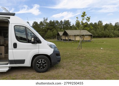 White camper van with open side door parked beside canvas glamping tents on a grassy campsite surrounded by pine trees, evoking travel, outdoor adventure and relaxed summer vacation vibes.