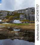 A white camper van is parked on a winding road through the rugged mountains of Norway. The van is reflected in a small, clear pool of water.