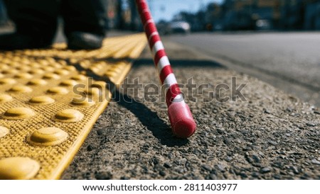 A white cane with red stripes rests on a yellow tactile paving surface next to a street. The cane is angled towards the viewer, with the tip in focus.