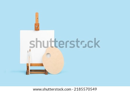 White canvas with copy space on a wooden easel and artist palette on a light blue background