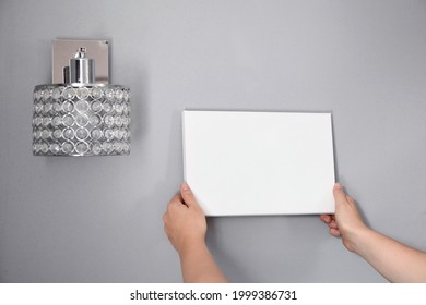 White Canvas In Female Hands With Gray Wall Background. Woman Hanging Blank Picture Mockup On Wall With Sconce. Wall Decor. Canvas Stretched On Frame