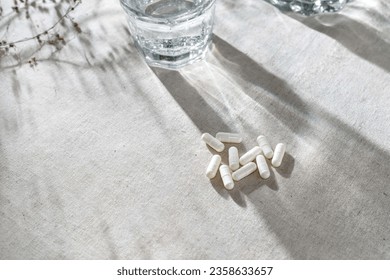 White capsules, glass with water and dried grass on neutral beige linen tablecloth with aesthetic light shadows.