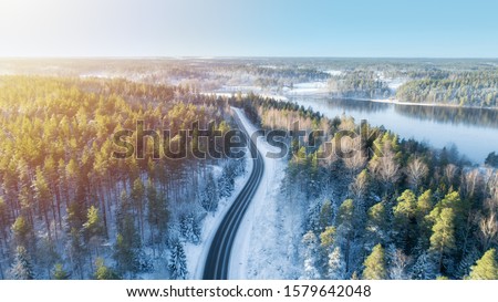 White car drives empty road running along the beautiful blue lake in the cold Finnish winter. Tourists on road trip cruising through the idyllic snow covered countryside and woods.