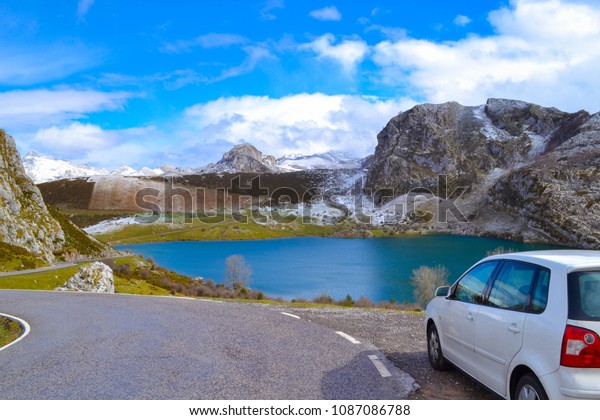 White car in Enol Lake in Picos de\
Europa, Asturias, Spain. Beautiful view of a blue and clear lake in\
middle of mountains, with a car and road at the\
front