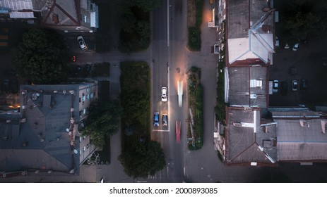 A White Car With Lights On Drives Along The City's Evening Street Among Houses And Trees - An Overhead Drone Shot.