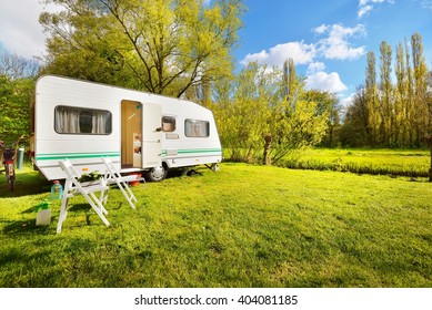 White Caravan Trailer On A Green Lawn In A Camping Site. Sunny Day. Spring Landscape. Europe. Lifestyle, Travel, Ecotourism, Road Trip, Journey, Vacations,  Recreation, Transportation, RV, Motorhome