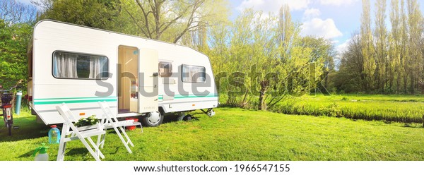 White caravan trailer on a green lawn in a\
camping site. Sunny day. Spring landscape. Europe. Lifestyle,\
travel, ecotourism, road trip, journey, vacations, recreation,\
transportation, RV,\
motorhome