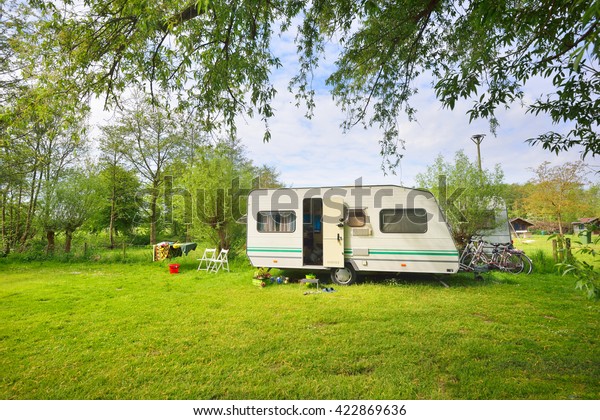 White caravan trailer on a green lawn in a\
camping site. Sunny day. Spring landscape. Europe. Lifestyle,\
travel, ecotourism, road trip, journey, vacations, recreation,\
transportation, RV,\
motorhome