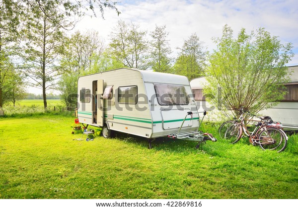 White caravan trailer on a green lawn in a\
camping site. Sunny day. Spring landscape. Europe. Lifestyle,\
travel, ecotourism, road trip, journey, vacations,  recreation,\
transportation, RV,\
motorhome