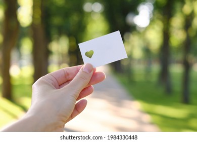 White Card With A Cut Out Heart In The Hands Of A Girl In The Rays Of The Sun. Close-up Postcard. Form For Congratulations. Romantic Invitation. Mockup. Empty Space For A Message Or Invitation