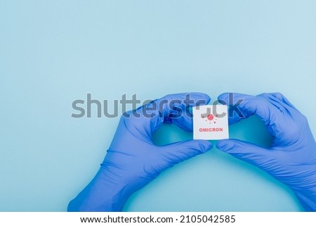 white card with red omicron lettering and bacteria icon in hands of doctor in latex gloves on blue, cropped view