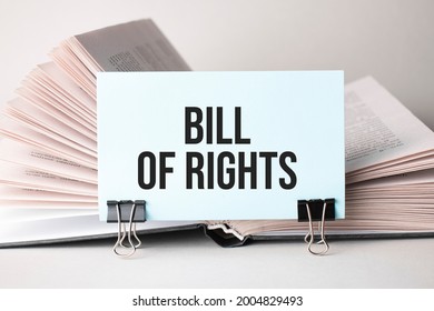 A White Card With The Text Bill Of Rights Stands On A Clip For Papers On The Table Against The Background Of Books. Defocus