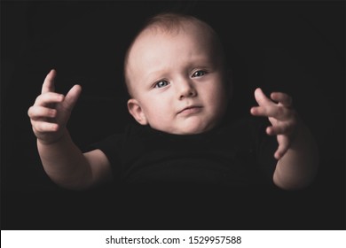 White Caucasian Beautiful Blue Eyed Baby Boy Is Showing His Emotions With Hands Reaching Towards The Viewer. Shallow Depth Of Field, Black T-shirt On Black Isolated Background. 