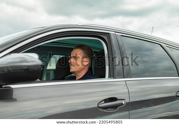 White Caucasian\
confident man is sitting on the drivers side of a gray black modern\
car and is looking straight forward at the road ahead. There are no\
trademarks in the shot.