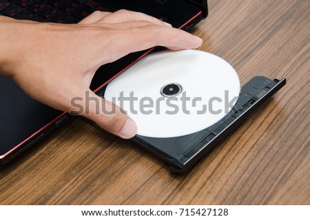 White CD or DVD ROM inserted in laptop computer on wooden texture background and human hand