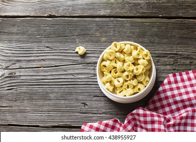 White Ceramic Bowl Full Of Italian Pasta Tortellini To Cooking On A Dark Wooden Background With Structural Tissue In A Red White Checkered.