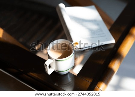 White ceramic coffee mug with light brown beverage beside open book on wooden table under natural sunlight