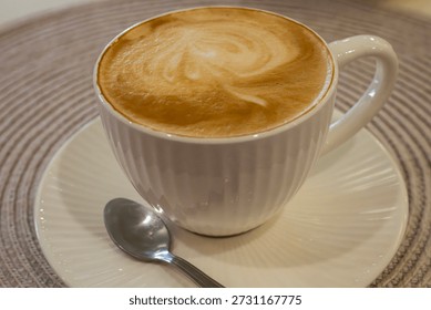 a white ceramic cup filled with a frothy cappuccino featuring latte art, resting on a matching saucer with a silver spoon on a textured beige placemat.