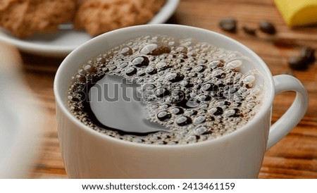 White ceramic cup with freshly brewed aromatic coffee and foam bubbles. A cup with a coffee or tea drink stands on the table near cookies and coffee beans. Morning ritual, breakfast concept. Close up.