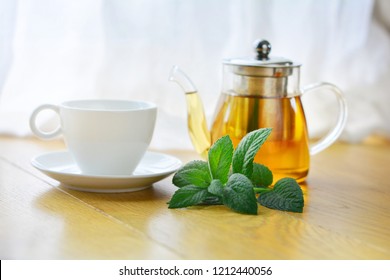 White ceramic cup and glass teapot of herbal tea with fresh mint twig on wooden table