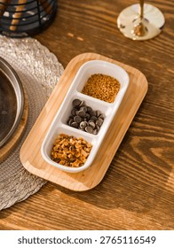 A white ceramic divided server filled with chocolate chips and spices is placed on a wooden tabletop in soft diffused light. A detailed shot reveals food ingredients arranged in separate sections under indoor daylight, with soft natural shadows.