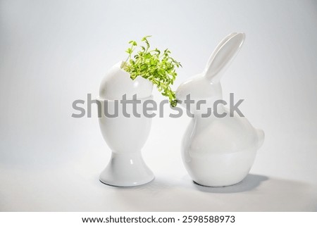 White ceramic Easter bunny eating cress from an eggshell in an egg cup, light gray background, copy space, selected focus, narrow depth of field