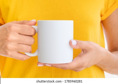 White Ceramic Mug Mockup. Woman Wears Yellow T-shirt Holding A Warm Cup Of Coffee. Copy Space For Your Logo