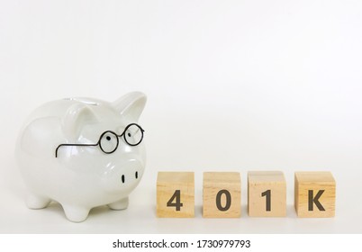 White ceramic piggy bank wearing reading glasses with wooden blocks spelling “401K” on white background. Concept for money savings plan for retirement, aged society, financial accounting