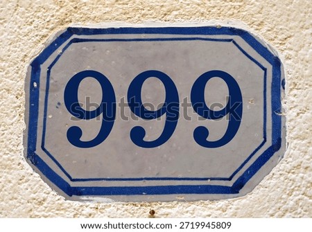 A white ceramic plate mounted on a textured wall showing a vintage blue house number 999 (nine hundred ninety nine)