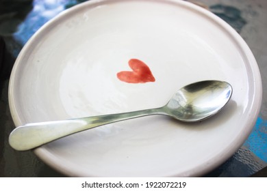 A white ceramic saucer with a picture of a red heart and a silver teaspoon on the surface of the plate. Clean utensils. High quality photo.