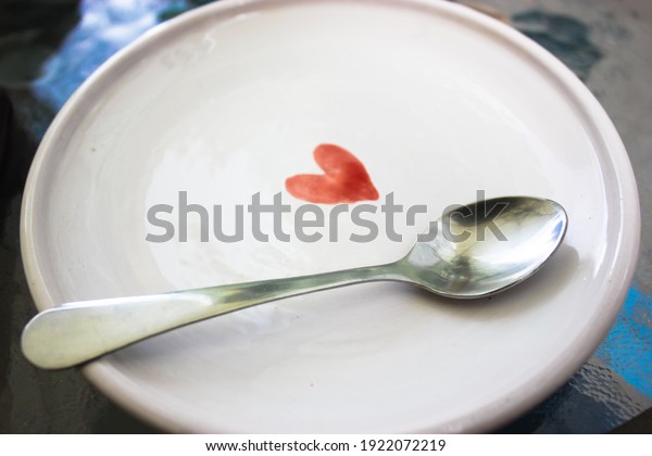 A white ceramic saucer with a picture of a red\
heart and a silver teaspoon on the surface of the plate. Clean\
utensils. High quality photo.