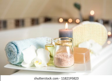 White Ceramic Tray With Home Spa Supplies In Home Bathroom For Relaxing Rituals. Candlelight, Salt Soap Bar, Bath Salt In Jar, Massage, Bath Oil In Bottle, Blue Rolled Towel, Natural Sponge. 