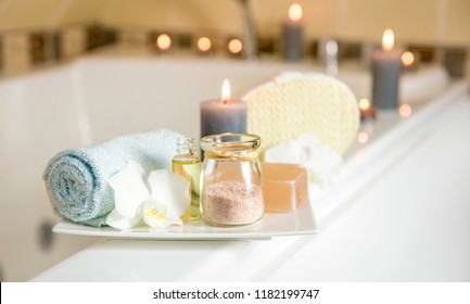 White Ceramic Tray With Home Spa Supplies In Home Bathroom For Relaxing Rituals. Candlelight, Salt Soap Bar, Bath Salt In Jar, Massage, Bath Oil In Bottle, Blue Rolled Towel, Natural Sponge. 