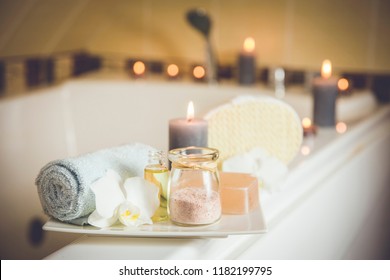 White ceramic tray with home spa supplies in home bathroom for relaxing rituals. Candlelight, salt soap bar, bath salt in jar, massage, bath oil in bottle, blue rolled towel, natural sponge. 