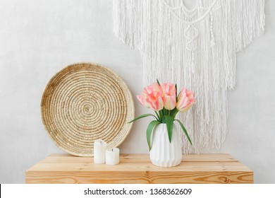White ceramic vase, bouquet of pink tulips flowers, wicker tray, candles, macrame wall hanging decoration on a wooden table or shelf on a background of light gray wall. Spring boho home interior decor
