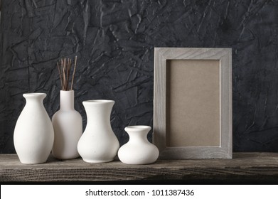 White Ceramic Vases And Wood Photoframe On Distressed Wooden Shelf Against Rough Plaster Black Wall. Home Decor.