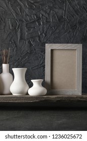 White Ceramic Vases And Wood Photoframe On Distressed Wooden Shelf Against Rough Plaster Black Wall. Home Decor.